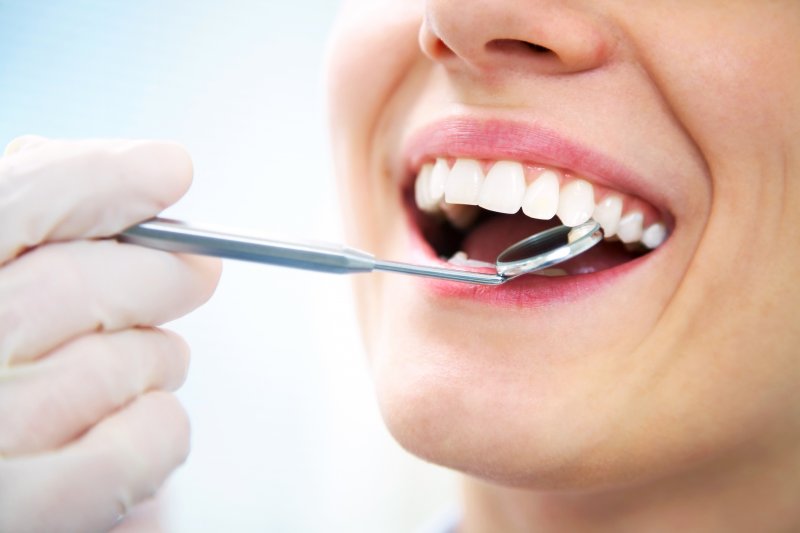 a dentist using a handheld dental mirror to look inside a women’s mouth who is smiling